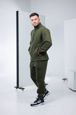 Kendine güvenen bir adam minimalist bir stüdyoda şık bir zeytin yeşili kıyafet ve spor ayakkabılar giyiyor. Parlak çevre onun rahat ama modaya uygun görünümünü vurguluyor..