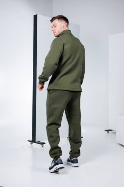 Kendine güvenen bir model, gösterişli, minimalist bir stüdyoda zeytin yeşili bir eşofman giyiyor. Parlak ışık, kıyafetin modern tasarımı ve dokusunu vurguluyor..