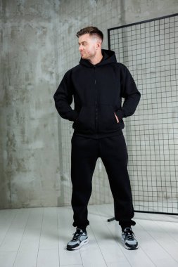 Erkek bir model, gri bir duvara ve basit bir şebeke arkaplanına dayanan parlak, siyah bir atletizm elbisesinin içinde güvenle duruyor. Görünüşü çağdaş şehir modasını temsil ediyor..