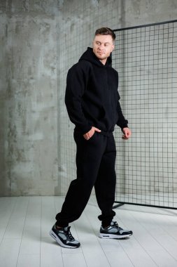 Siyah bir giysi giymiş, minimalist bir stüdyoda duran genç bir adam. Kendine güveni tam görünüyor, rahat ama şık bir hava yayıyor..