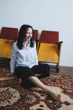 Nazik ışıkla dolu sakin bir kapalı alanda, genç bir kadın zarif, renkli bir halının üzerinde oturur, eski sandalyelerle çevrili, huzur ve sakinlik saçar.