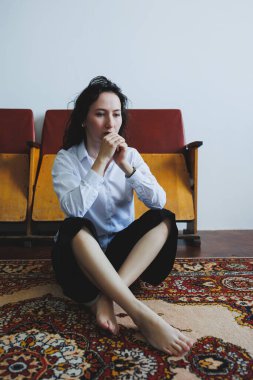 Süslü bir halıya çıplak ayakla oturan bir kadın, sade bir beyaz gömlek ve parlak siyah pantolon giyerek rahatlama ve içgözlem için ideal sakin bir atmosfer yaratıyor.