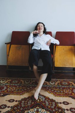 Şık beyaz gömlekli ve siyah pantolonlu rahat bir kadın şık sandalyelerde zarif bir şekilde uzanır, güzel bir halıyla çevrili, rahat bir atmosferde zarafet saçar.