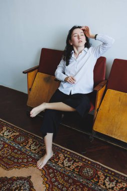 Beyaz gömlekli ve siyah pantolonlu genç bir kadın klasik bir sandalyede çıplak ayakla oturur, rahat ama düşünceli bir titreşim yayar.