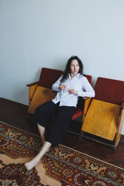 Sakin bir iç mekan ortamında, bir birey rahat giysiler giyerek tahtadan sandalyelere rahatça oturur. Halı ve duvarların ılık tonları samimi bir ortam yaratıyor..