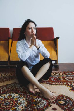 Kıvırcık saçlı bir kadın derin düşüncelere dalmış renkli bir halıda bacak bacak üstüne atmış oturuyor. Beyaz bir gömlek ve siyah bir etek giyiyor. Eski tahta sandalyelerle çevrili. Huzurlu bir atmosfer yaratıyor..