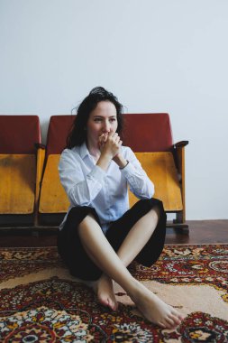 Siyah saçlı genç bir kadın canlı bir halıda bacak bacak üstüne atarak oturuyor. Çıplak ayaklarıyla yumuşak kumaşa dokunuyor. Düşünceli bir şekilde ileri bakıyor, sakin bir odada kırsal sandalyelerle çevrili..