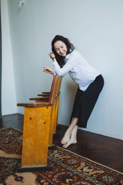 Genç bir kadın şakacı bir şekilde tahta sandalyeye yaslanıyor, yalın ayak ve parlak bir odada hafifçe gülümsüyor. Renkli halı, dingin atmosfere sıcaklık katıyor..