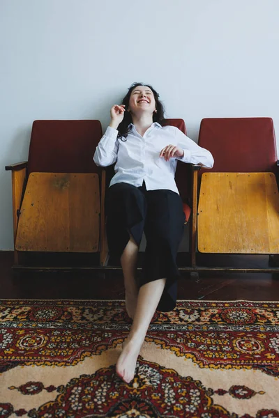 Sıcak, davetkar bir odada, bir kadın basit bir beyaz gömlek ve siyah pantolon giyer, retro bir bankta oturur. Neşeli bir şekilde gülüyor, rahatlatıcı enerji ve konforla dolu neşeli bir atmosfer yaratıyor..