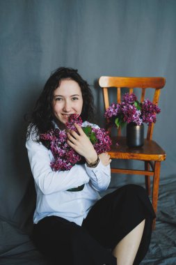 Neşeli bir kadın elinde çiçekli leylak buketi, kırsal bir sandalye ve vazoyla yerde oturuyor. Renk ve yaşamla dolu sıcak ve davetkar bir atmosfer yaratıyor..