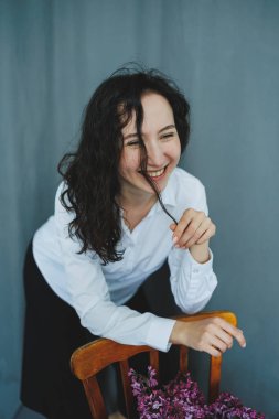 Sakin, minimalist bir alanda kıvırcık saçlı genç bir kadın ahşap bir sandalyedeki çiçeklere neşe ve güzellikle dolu neşeli bir anı yakalıyor..