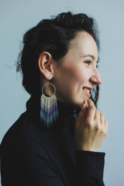 Neşeli bir kadın çenesine dokunurken eşsiz küpelerini sergiliyor. Açık mavi arka plan, bu samimi ortamda onun canlı görünüşünü arttırıyor..