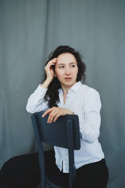 Kıvırcık saçlı bir kadın düşünceli bir şekilde sandalyeye oturur, eli başının üzerinde dinlenir, yansımayı davet eden basit gri bir ortamda sakin bir atmosfer yaratır..