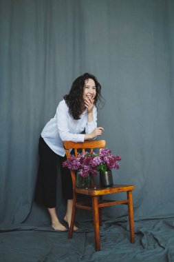 Neşeli bir kadın basit bir ahşap sandalyenin yanında durur, üzerine konulmuş canlı çiçeklerle mutlu bir şekilde etkileşim içindedir. Pürüzsüz gri arka plan, neşeli ifadesi için sakin bir atmosfer yaratıyor..