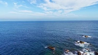 Yumuşak dalgalar, sakin mavi bir okyanusta yuvarlanarak kıyıdaki pürüzsüz kayalara ulaşır. Yumuşak bulutlu canlı bir gökyüzü huzur ve yansımaya güzellik katar..