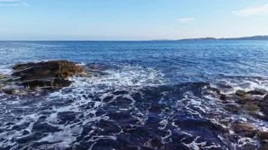 Güneş dingin okyanusu aydınlatırken dalgalar yavaşça karanlık kayalara vurur. Mavi gökyüzü tepemizde uzanıyor, sahilde huzurlu bir atmosfer yaratıyor..