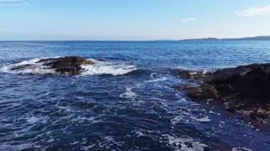 Dalgalar açık mavi gökyüzünün altında kıyı kayalarına hafifçe çarpar. Uzak tepeler ufukta belirdi, güneşli bir günde sakin ve sakin bir atmosfer yarattı..