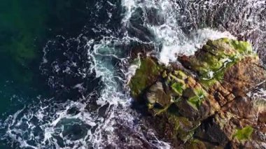 Yumuşak dalgalar, yeşil yosunlarla kaplı sivri kayaların üzerinden süzülüyor. Derin mavi su zengin dokularla çelişiyor, alacakaranlıkta doğanın sakin güzelliğini yakalıyor..