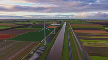 Rüzgâr türbinleri, yeşil ve kahverengi renkli canlı tarlalarla çevrili sakin bir kanalın yanında dururlar. Değişen bulutlar, öğleden sonra ışığı altında güzel bir duvar halısı oluşturur..