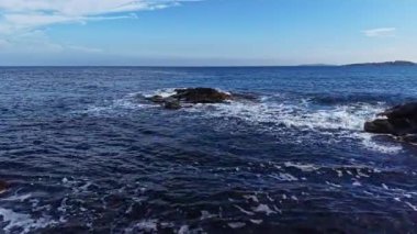 Gün boyunca sakin bir kıyı sahnesini aydınlatan, berrak mavi sularda parlayan güneş ışığı gibi yumuşak dalgalar kayalık oluşumlar üzerinde yuvarlanır..