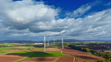 Uzun rüzgar türbinleri, rüzgar enerjisini kullanırken zarifçe dönen yeşil tepelerde ve kahverengi tarlalarda gururla durur. Manzara parlak renklerle dolu, doğanın güzelliğini gözler önüne seriyor.