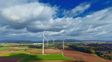 Uçsuz bucaksız yeşil alanlar kilometrelerce uzanıyor, yüksek rüzgar türbinleri yavaşça dönüyor. Beyaz bulutlar parlak mavi gökyüzünde yüzerler, doğanın güzelliğindeki sürdürülebilir enerjiyi temsil ederler..