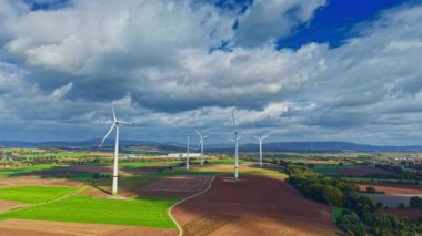 Uzun rüzgar türbinleri, kabarık bulutlarla dolu açık mavi bir gökyüzünün altında zarif bir şekilde döner. Alanlar uyumlu renklerde uzanıyor, dinamik ve dinamik bir sahne sunuyor.