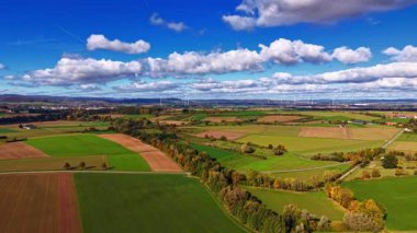 Berrak bir gökyüzünün altında rüzgar türbinleri yavaşça dönerken, zengin yeşil ve kahverengi alanlardan oluşan esneme alanları manzarayı süsler. Bu sakin kırsal alanda doğa gelişiyor..