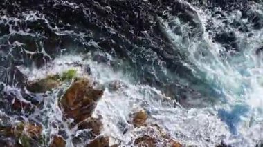 Güçlü dalgalar sivri kayalarla çarpışarak havaya su püskürtüyor. Güneş ışığı okyanusta dans eder, çarpan dalgaların ortasındaki mavi ve kahverenginin canlı kontrastını vurgular..