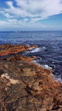Dalgalar kıyı şeridi boyunca uzanan engebeli kayalara hafifçe vururken, güneş tepemizde parlak bir şekilde parlarken, sakin ve canlı bir kıyı manzarası yaratıyor..