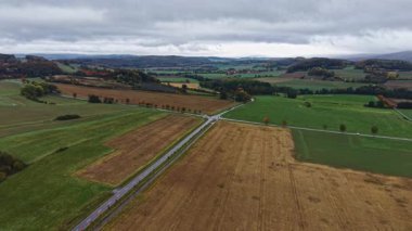 Huzurlu bir kavşak canlı yeşil tarlaları ve kasvetli bir gökyüzü altında altın-kahverengi tarım arazilerini ayırır. Sonbahar renkleri engin, açık kırsal alanda uyum içinde harmanlanır..