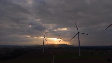 Güçlü rüzgar türbinleri gün batımının solan ışığını yakalıyor, gölgeleri yavaş yavaş karanlık bulutlara karşı dönüyor, sakin bir akşamda doğa ve teknolojiyi uyum içinde sergiliyor..