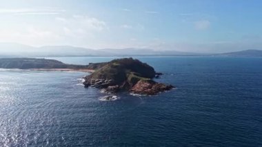 Sakin sular ve uzak dağlarla çevrili kayalık bir ada, parlak gökyüzünün altında huzurlu bir manzara yaratıyor..