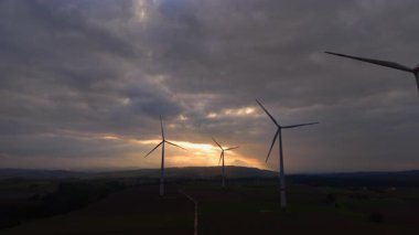 Günbatımı, pitoresk bir manzarada yer alan rüzgar türbinlerine altın bir parıltı saçar. Gökyüzü bulutlarla dolu ve temiz enerji üretimi için doğa ile uyum içinde çarpıcı bir zemin oluşturuyor..