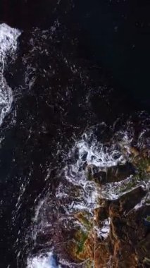 Beyaz uçlu dalgalar kıyı şeridi boyunca dalgalanan engebeli kayalara karşı, karanlık sular ve güneşli sahil arasında çarpıcı bir kontrastlık yaratarak, doğanın öğleden sonra ışığı altındaki ham güzelliğini vurguluyor..