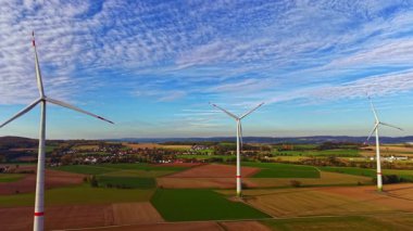 Uzun rüzgar türbinleri yemyeşil bir arazide dururlar ve temiz enerji üretirler. Gökyüzü kabarık bulutlarla boyanmış, bölgenin doğal güzelliğini vurguluyor..