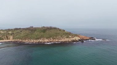 Serene sahili yeşil ve kayalık kıyıları olan küçük bir adayı gösteriyor. Yumuşak dalgalar plajda sisli bir gökyüzünün altında, barışçıl bir atmosfer yaratır..
