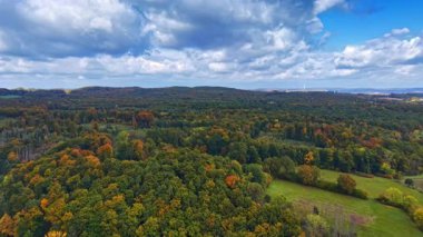 Renkli sonbahar yaprakları yuvarlanan tepeleri örterek çarpıcı bir manzara yaratır. Parlak güneş ışığı kabarık bulutların arasından süzülür, doğanın canlı tonlarını aydınlatır..