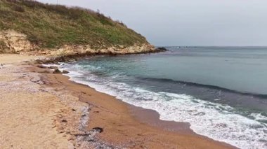 Yumuşak dalgalar bulutlar gökyüzünü örterken kumlu sahili nazikçe öpüyor. Bereketli yeşil bir yamaç sahilin ötesine yükseliyor. Sabahın ilk ışıklarıyla deniz kenarında huzurlu bir kaçış sunuyor..