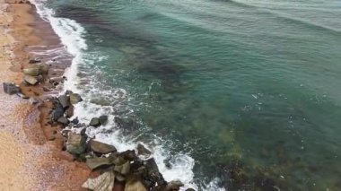 Güneş batarken dalgalar engebeli bir kıyı şeridine yuvarlanıyor. Su mavi ve yeşilin tonlarında parıldıyor. Sahilin kenarında sessiz bir yansıma yaratıyor..