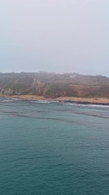 Sakin sular sisli bir günün sessiz ışığını yansıtırken, kayalıklar kıyı boyunca görkemli bir şekilde yükselirken, gözlerden uzak bir plaj ve yemyeşil bitki örtüsünün ardında bekliyor..