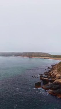 Güneş sisli bir kıyı şeridinde yükselirken dalgalar yavaşça karanlık kayalara çarpar. Manzarada engebeli bir kıyı ve yeşillikle kaplı uzak bir kara parçası var..