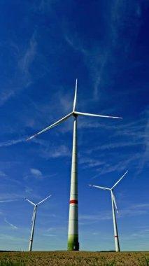 Mavi gökyüzüne karşı uzun beyaz yel değirmenleri. Rüzgar türbinleri dönüyor ve alternatif enerji üretiyor. Avrupa 'nın kırsal kesimlerinde bir tarlada yel değirmenleri.