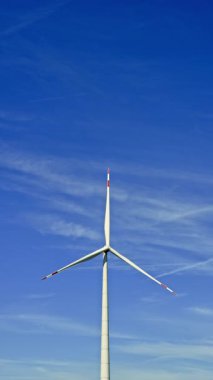 Mavi gökyüzüne karşı büyük bir rüzgar türbini dönüyor. Yel değirmenlerinin yeşil enerji üretimi kırsal bir alana yerleştirildi.