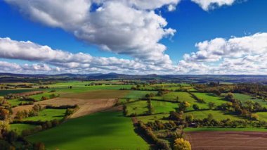 Ağaçlarla çevrili arazide altın ve yeşil tarlalar uzanırken, tüylü bulutlar parlak mavi bir gökyüzünde süzülüyor, kırsal kesimde huzurlu bir sonbahar gününün güzelliğini yakalıyor..