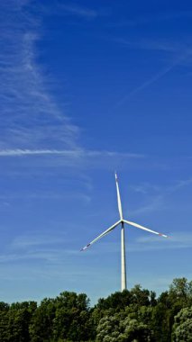 Mavi gökyüzüne karşı büyük bir rüzgar türbini dönüyor. Yel değirmenlerinin yeşil enerji üretimi kırsal bir alana yerleştirildi.