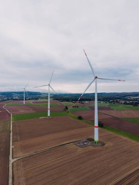 Uzun rüzgar türbinleri, sakin bir tarımsal alanda yavaşça döner. Alanlar, doğanın güzelliğini ve sürdürülebilir enerji kaynaklarına olan bağlılığını gösteren her yöne doğru uzanıyor..