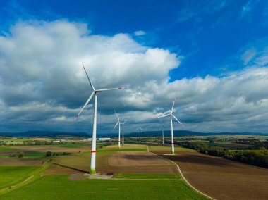 Dev rüzgar türbinleri, zengin kahverengi tarlalar ve canlı yeşilliklerle dolu engin bir tarım arazisinde zarifçe dönerler. Kabarık bulutlar gökyüzünde sürükleniyor, güzel bir sahne yaratıyor..