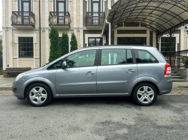 Gümüş renkli kompakt bir araba çok katlı güzel bir binanın yanına güzelce park edilmiştir. Çevre alanı huzur dolu, ağaçlar sahneye doğanın dokunuşunu katıyor..