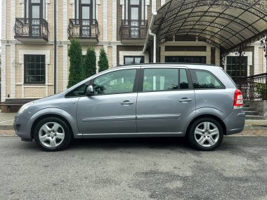 Parlak gümüş renkli bir minivan zarif taş detaylar ve yeşil çalılarla güzel bir mimari binanın yanına park edilmiş ve öğleden sonra ışığında sakin bir şehir manzarası yaratıyor..